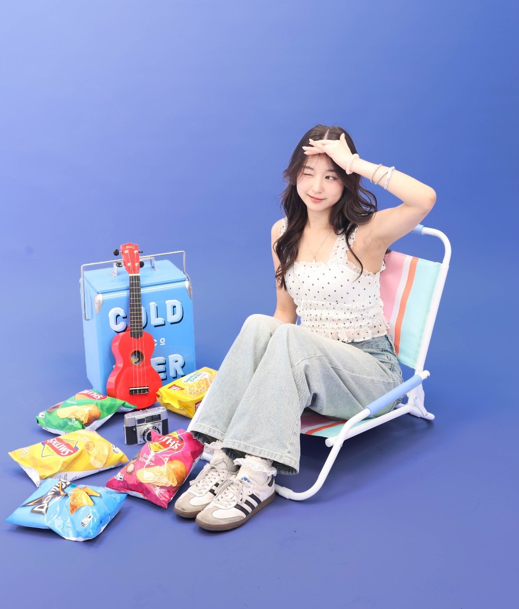 A person winking from a striped beach chair on a blue set, surrounded by chip packets, a red ukulele, a cooler and a vintage camera