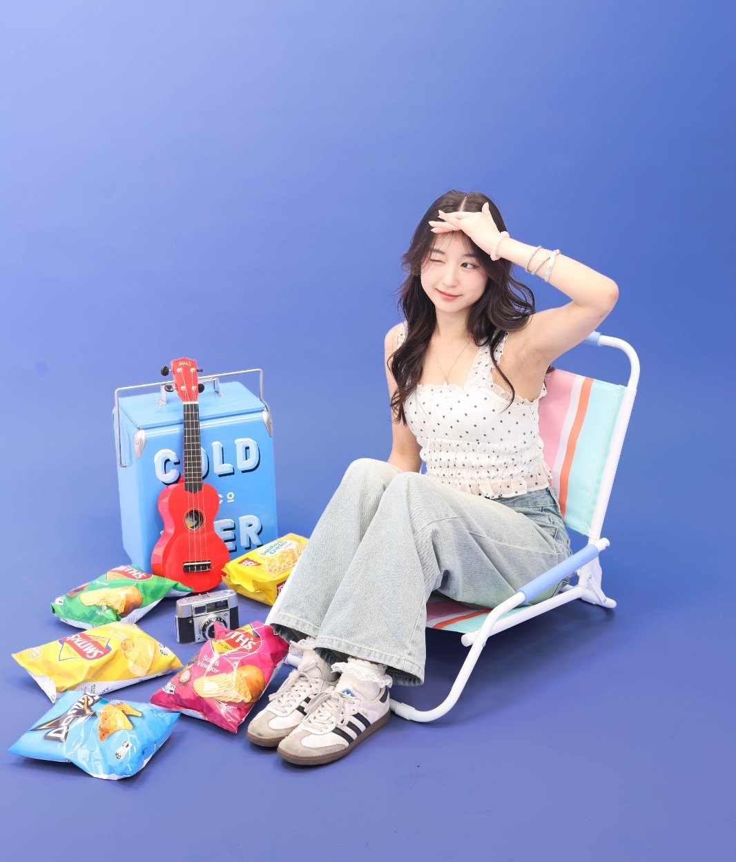 A person winking from a striped beach chair on a blue set, surrounded by chip packets, a red ukulele, a cooler and a vintage camera
